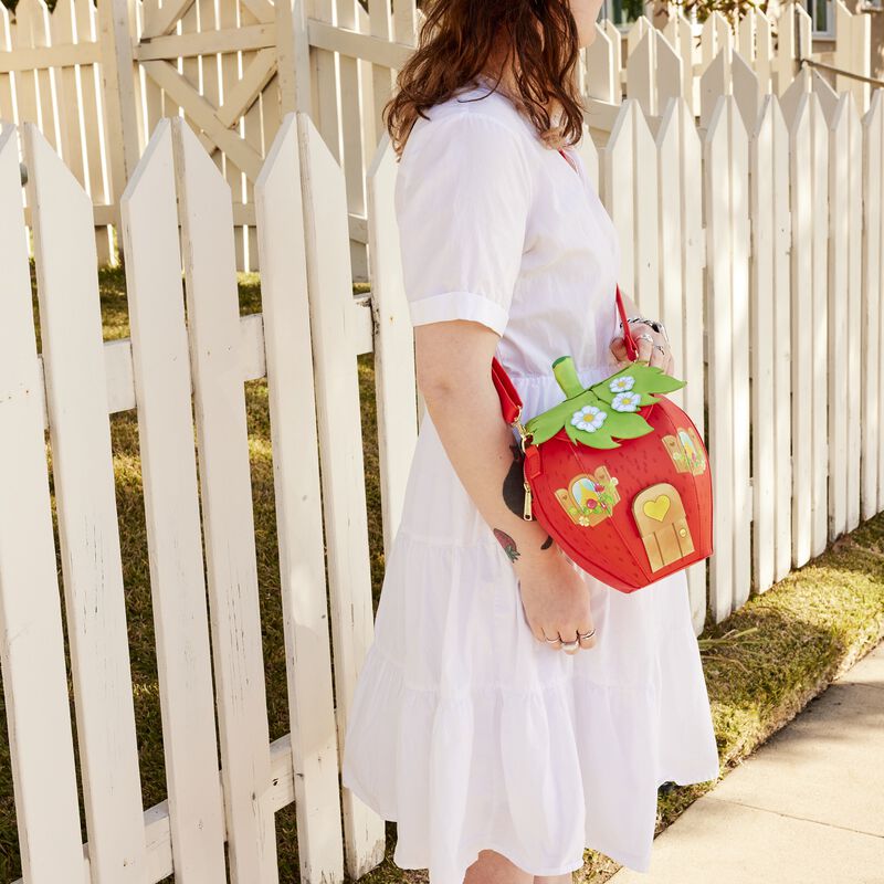 Woman wearing a white dress, standing in front of a white picket fence, wearing our figural Strawberry Shortcake Strawberry House Crossbody Bag